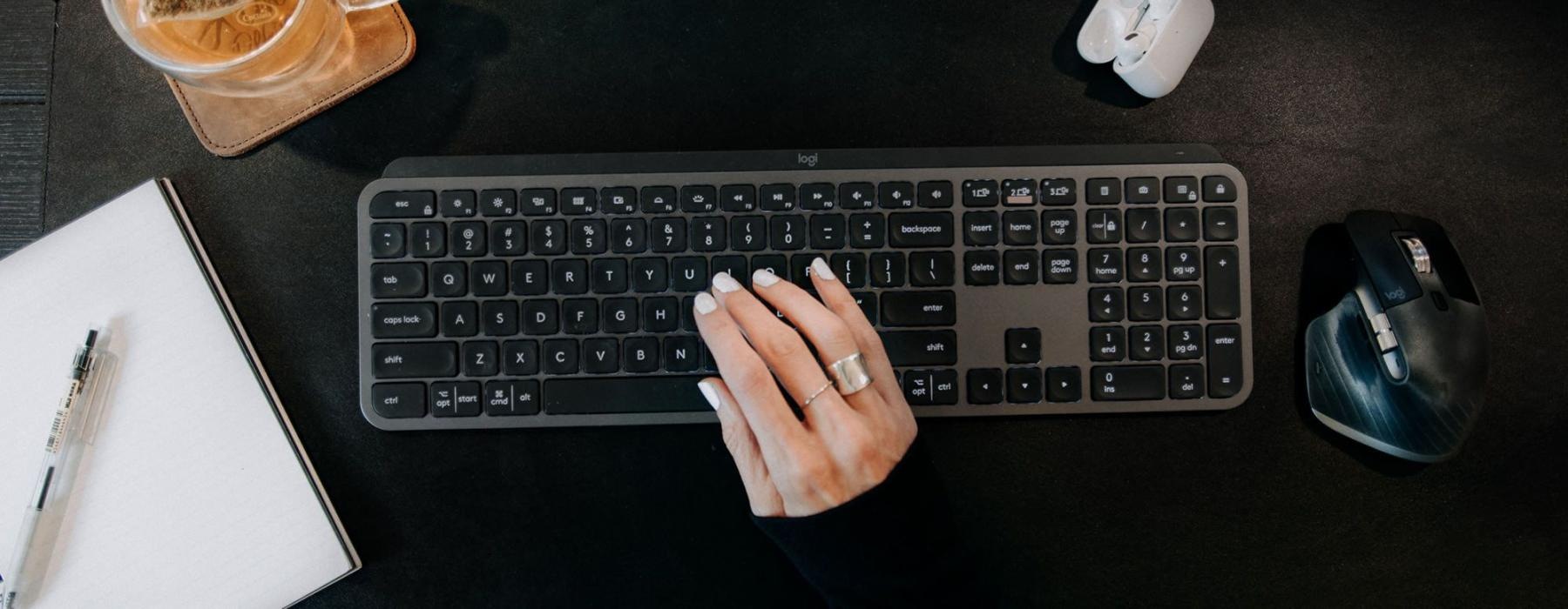 woman's hand on a keyboard surrounded by office items and a cup of tea on a coaster