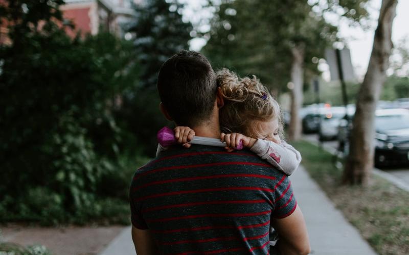 tired little girl rests her head on her father's shoulder as he carries her down the city sidewalk